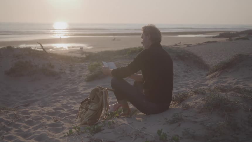 A person is sitting on a sand dune looking out at the ocean at sunset the person is holding a small notebook and appears to be writing or sketching