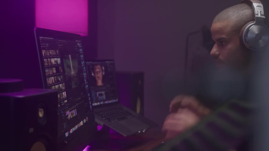 A male video editor is seen from the side sitting at a desk and putting on a pair of silver headphones he is working in a dark room with two monitors one of which shows a video editing program
