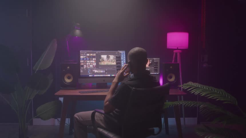 A man in a dark room sitting at a video editing desk watching a video