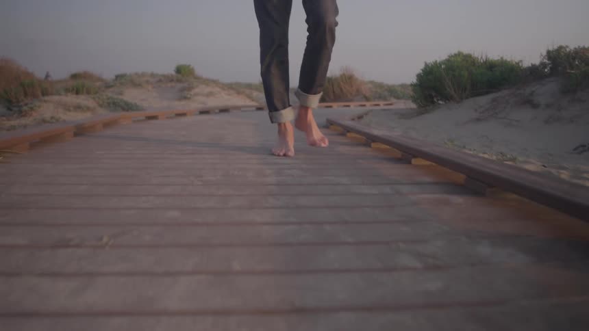 A person wearing gray pants with rolled up cuffs walks barefoot on a wooden boardwalk that leads through sandy dunes with dry green bushes