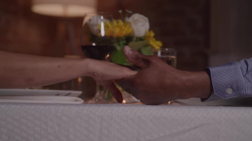 A woman and a man sit at a white tablecloth table with a candle and a vase of flowers reaching across the table to clasp each others hands
