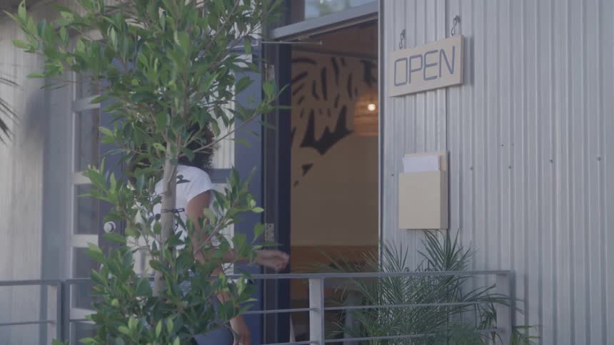 A woman with curly black hair stands on a small patio reaching up to hang a white sign with the word open on the outside wall of a building