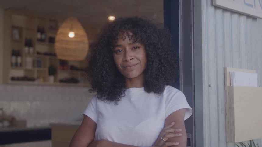 A woman with curly dark hair wearing a white t shirt stands with her arms crossed and smiles at the camera
