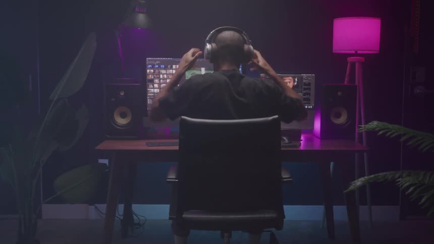 A person sits at a desk in a dimly lit room with purple and pink lighting putting on headphones and looking at multiple computer screens displaying video editing software