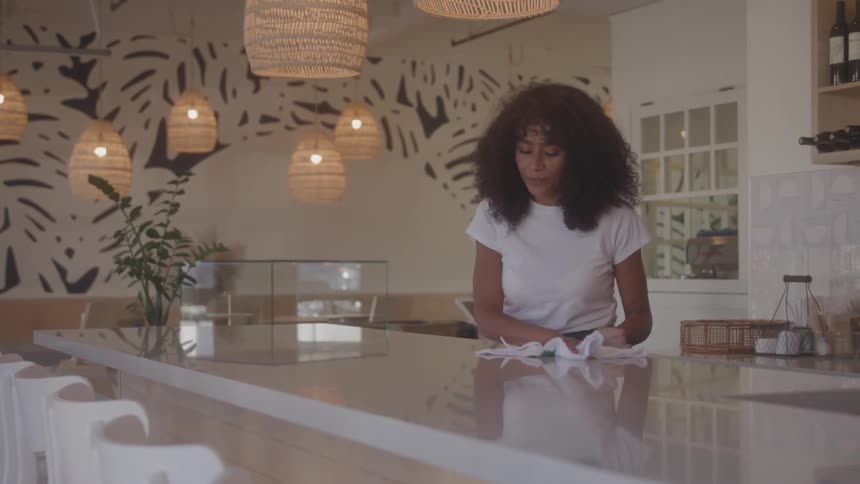 A shot of a woman with curly hair wiping a long reflective counter in a restaurant with a modern design