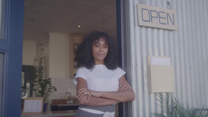 A smiling young woman stands cross armed at the door of a coffee shop next to the open sign