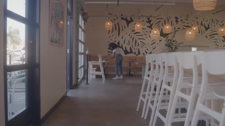 A waitress with curly black hair stands on a cafe where she sweeps the floor with a broom