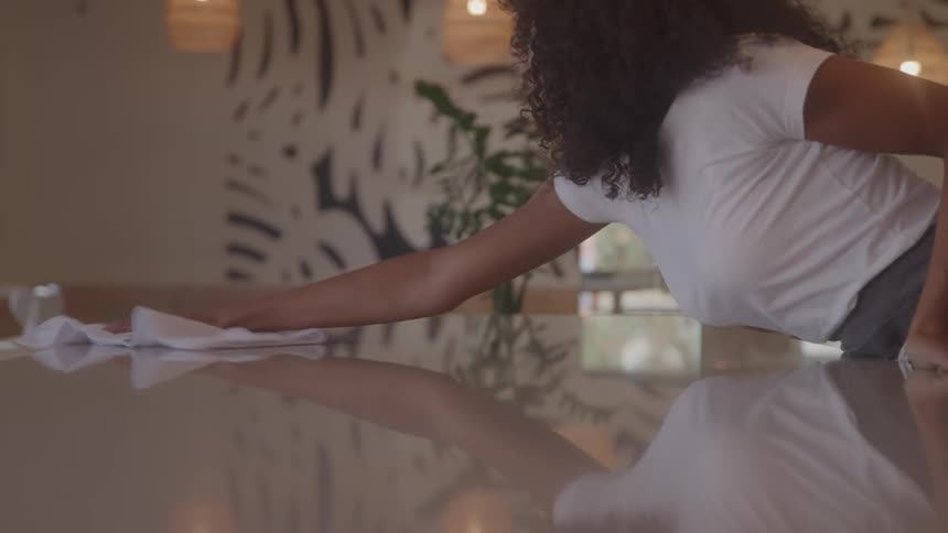 A shot of a woman with curly hair wiping a long reflective counter in a cafe setting