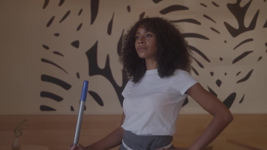 A woman with curly dark hair and a white tshirt stands in a room with a patterned wall holding a blue mop with her hands resting on her hips