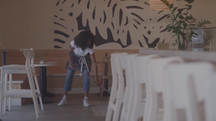 A woman with curly hair wearing a white shirt and apron sweeps the floor of a cafe