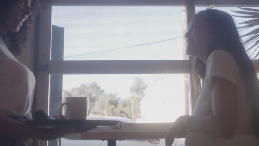 A female waiter served a young woman a cup of coffee as she sat at a windowside table inside a cafeteria