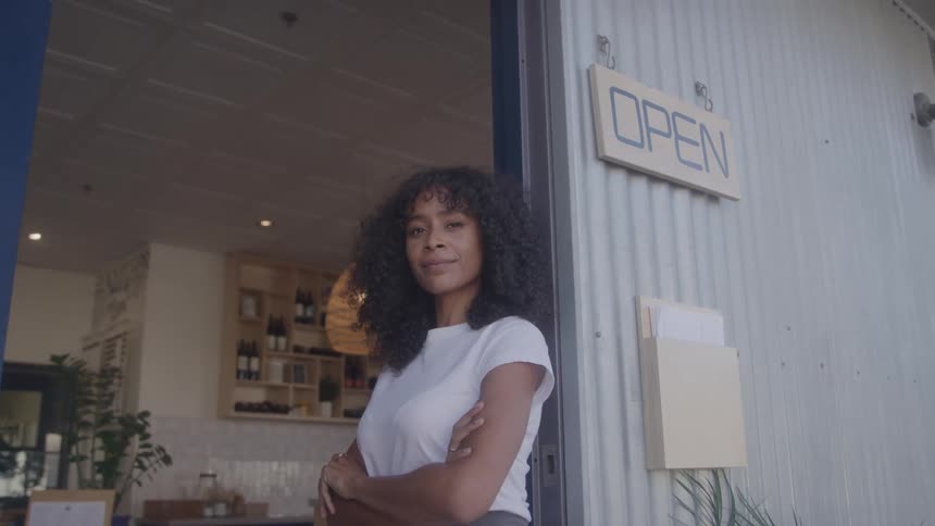 A woman with her arms crossed stands in the open doorway of a cafe