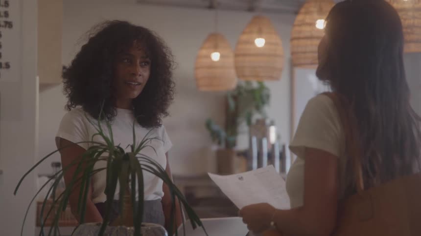 A young woman at the counter of a coffee shop gives a menu to a client and talks to her about it