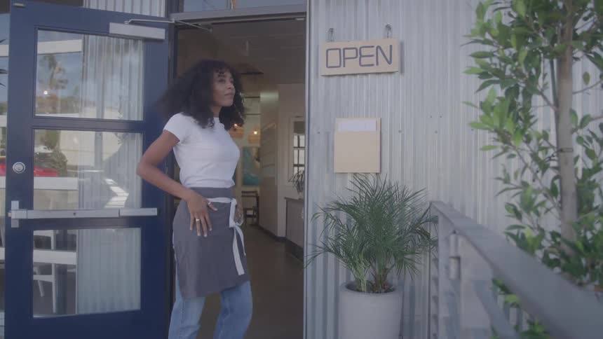 A woman with curly hair hangs an open sign on the door of a cafe before walking inside