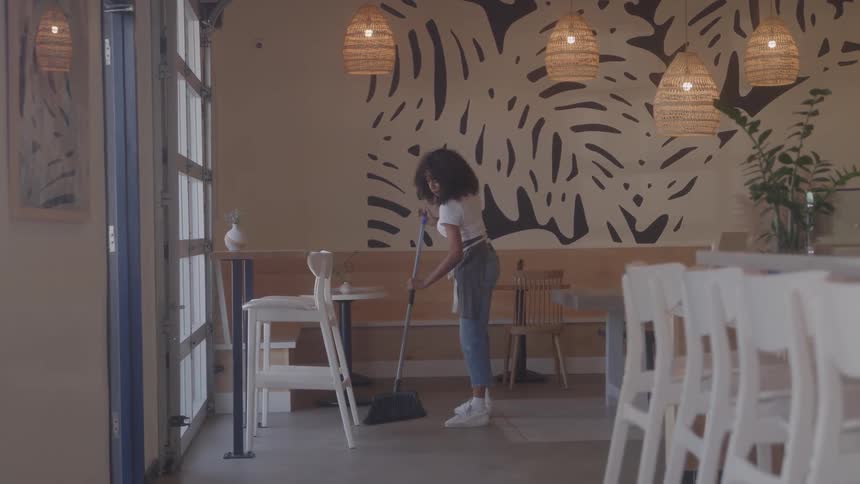 A young woman working in a closed coffee shop adjusting the chairs and sweeping the floor with a broom