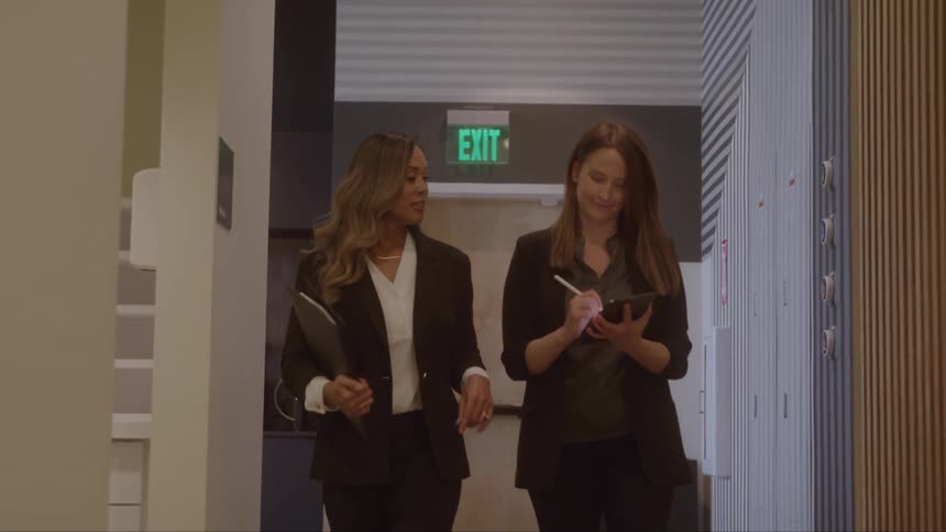 Two women in blazers talk while walking through an office hallway