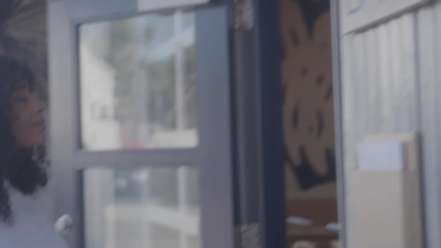 A woman with curly hair reaches up to a sign on a glass door flipping it from closed to open