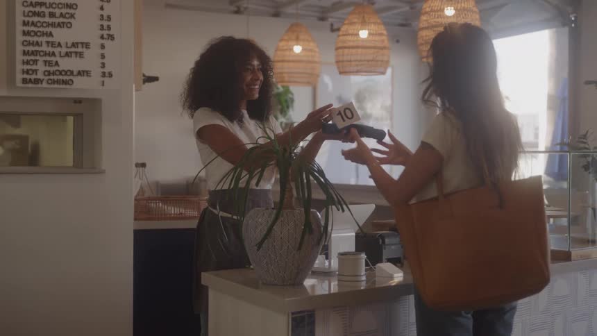 A barista with curly hair is at a counter smiling as she hands a card payment machine to a customer the customer is holding a large tote bag and is about to pay for their order