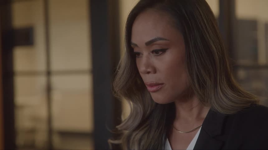 A young woman is shown in a closeup chestup shot she has long dark hair with blonde highlights and is wearing a black blazer and a gold necklace she looks down and then glances up appearing thoughtful or pensive