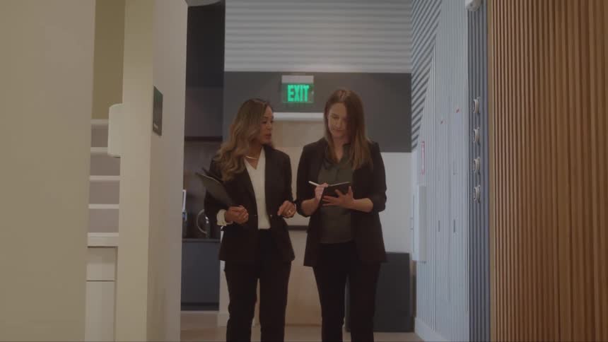 Two businesswomen walking down a hallway looking at a tablet as they discuss something