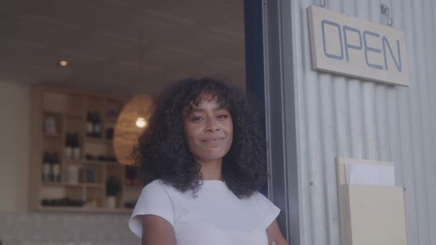 A smiling woman with dark curly hair stands confidently in front of a small cafe or shop an open sign is visible to the right of her indicating that the business is ready to welcome customers