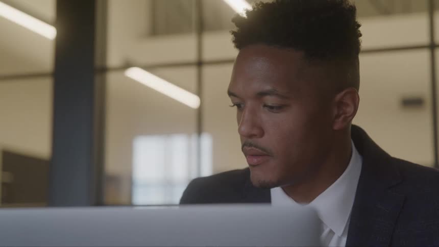 A closeup of a man in a professional suit with a serious expression focused on working on his laptop in a modern office