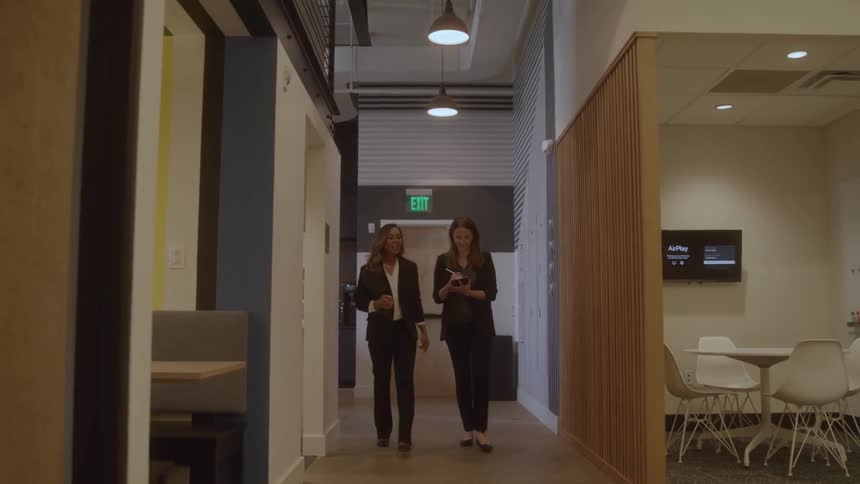 Two women walk down an open corridor in an office space both smiling one of them talking while the other takes notes