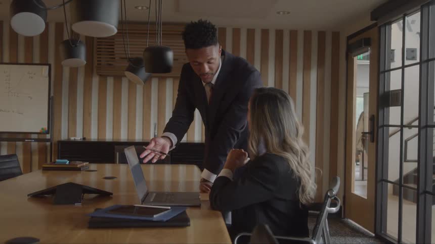 A man and a woman in an office with the woman sitting at a desk with an opened laptop in front of her and listening to the man standing beside her talking while pointing at the laptop