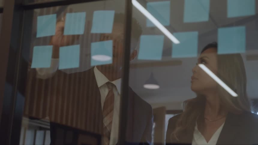 A man and a woman standing in an office next to a glass wall with postits analysing and discussing the content of the postits