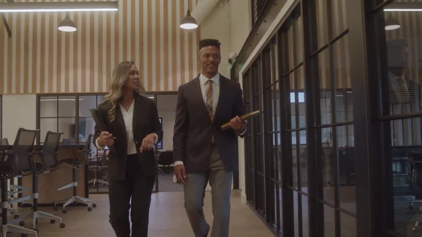Two colleagues in suits walk and talk in a modern office hallway