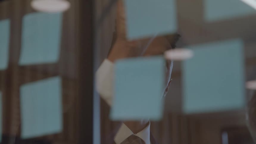 A businessman is shown through a transparent glass wall covered in blue sticky notes he is working with a female colleague and they appear to be discussing the notes on the wall