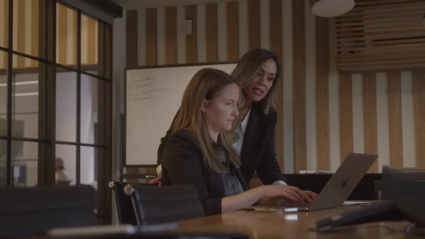Two businesswomen work together on a laptop inside a meeting room