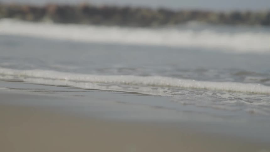 From a low groundlevel angle a thin line of white foam from a gentle wave washes ashore onto wet sand with larger outoffocus waves in the background