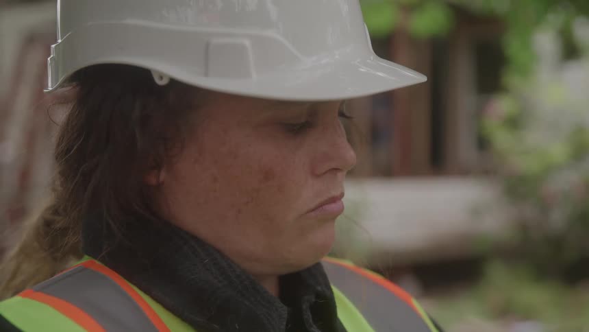 A person wearing a white hard hat and a highvisibility vest is shown in a closeup side profile shot they look down with a serious expression before slowly raising their gaze upwards