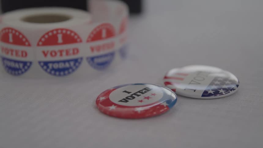 In a closeup shot a persons hand picks up a patriotic i voted button from a table next to a roll of voting stickers