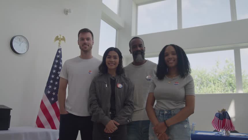 Four people two men and two women stand together in a white room with a large window and a clock on the wall as an american flag stands next to them