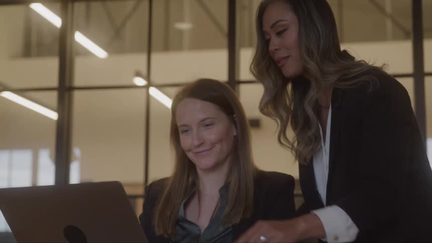 Two businesswomen collaborate while looking at a laptop in a modern office