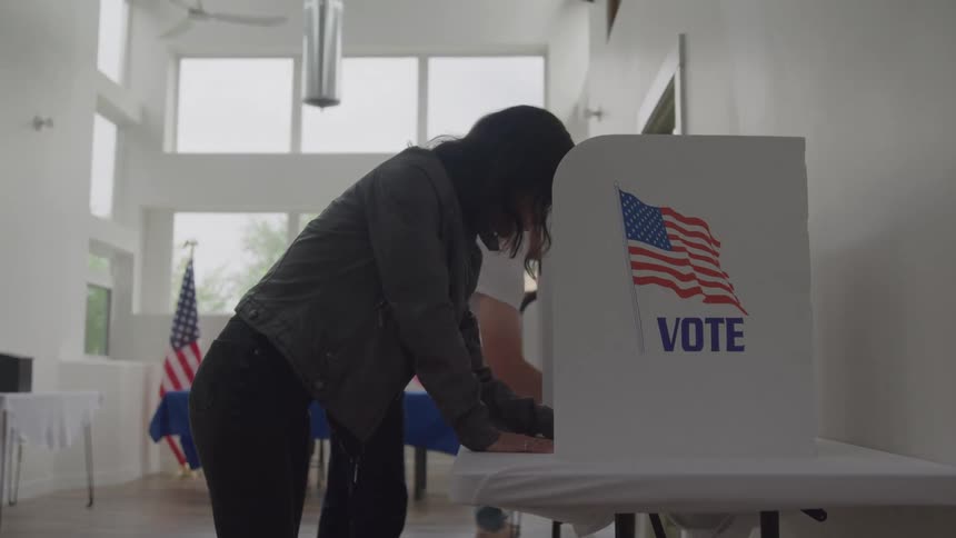 A person with dark hair wearing a gray jacket and black pants stands at a white voting booth with an american flag and the word vote on it filling out a paper ballot