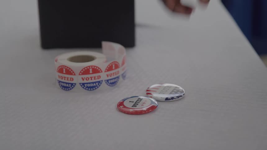 A closeup of a white table with a roll of i voted today stickers and two individual i voted pins and a human hand picking a pin