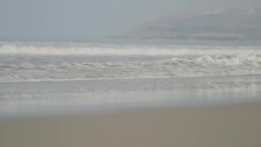In a static slowmotion shot lines of ocean waves with white foam break and roll toward a wet sandy shore on a hazy overcast day