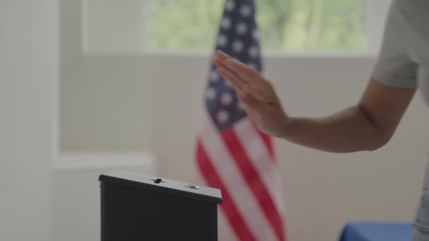 A persons arm places a voting ballot into a small black box on a table with an american flag visible in the background