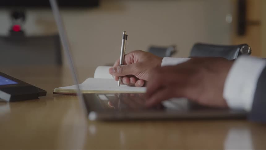 A lowangle closeup shot focuses on a persons hands as they type on a laptop keyboard they occasionally stop to pick up a pen and write notes in an open notebook