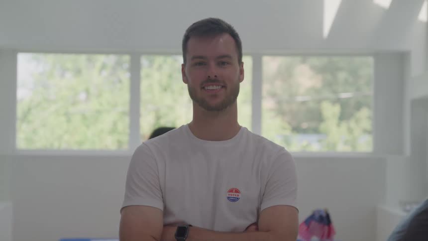 A man with a light complexion and a beard stands indoors with his arms crossed smiling at the camera and wearing a white tshirt with a blue and red sticker