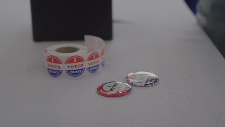 A persons hand picks up a red voter pin from a white table that also holds a roll of voted today stickers