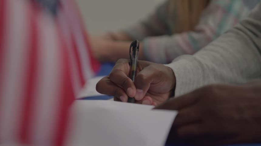 A black person writing with a pen on a piece of paper in a classroom at midday