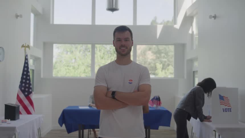 A male stands in the foreground with his arms crossed smiling at the camera in the background two other people are seen at a table registering to vote