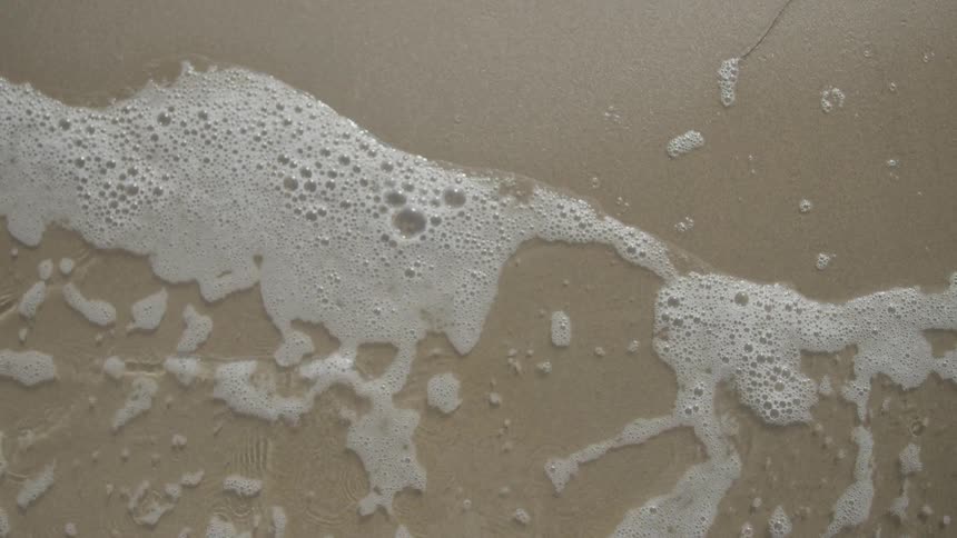 From a topdown perspective the foamy bubbling edge of a gentle ocean wave washes over and then recedes from the flat wet sand of a beach