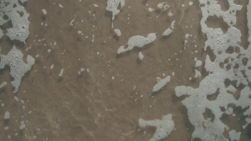 A macro shot from above shows the tide coming in over a sandy beach leaving a trail of seafoam the foam retreats back into the ocean revealing the wet sand underneath