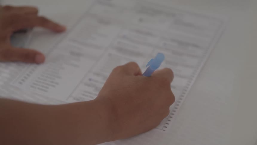 A persons hand holding a blue pen is seen from a closeup highangle shot the hand is marking choices on a paper form or ballot with a series of quick deliberate strokes