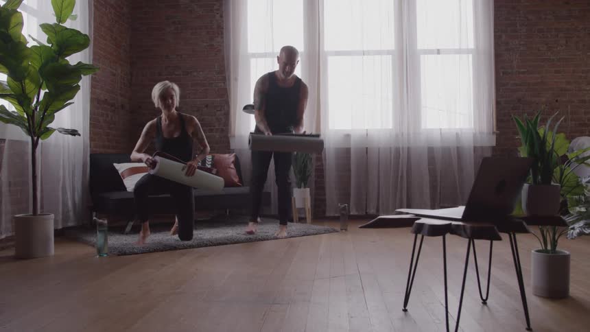 A middleaged man and a young woman walk into a living room at midday set down their water bottles on the floor and spread out their yoga mats ready to exercise